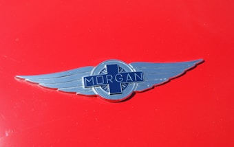 A silver Morgan car emblem with wings is centered on a glossy red background. The emblem features the word 'Morgan' across a blue cross-like design.