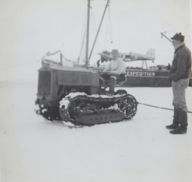 Two individuals are interacting near a snow-covered tracked vehicle in an icy environment. There is a ship with the visible text 'EXPEDITION' on its side, and an airplane is also present in the background. One person is seated on the tracked vehicle, while the other stands beside it.