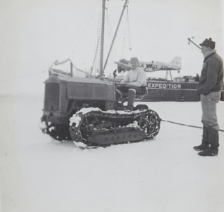 Two individuals are interacting near a snow-covered tracked vehicle in an icy environment. There is a ship with the visible text 'EXPEDITION' on its side, and an airplane is also present in the background. One person is seated on the tracked vehicle, while the other stands beside it.