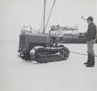 Two individuals are interacting near a snow-covered tracked vehicle in an icy environment. There is a ship with the visible text 'EXPEDITION' on its side, and an airplane is also present in the background. One person is seated on the tracked vehicle, while the other stands beside it.