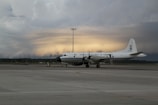 A large white aircraft with the inscription 'United States Dept. of Commerce' is parked on a tarmac. The sky in the background appears dramatic with layers of dark clouds and a faint glow of light, suggesting a sunrise or sunset.