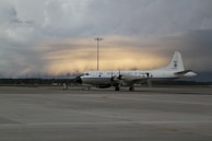 A large white aircraft with the inscription 'United States Dept. of Commerce' is parked on a tarmac. The sky in the background appears dramatic with layers of dark clouds and a faint glow of light, suggesting a sunrise or sunset.