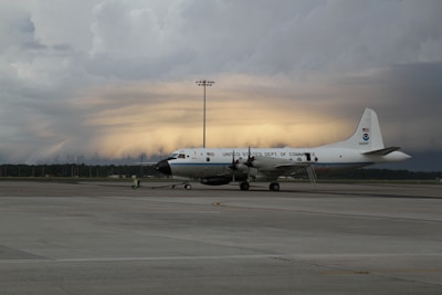 A large white aircraft with the inscription 'United States Dept. of Commerce' is parked on a tarmac. The sky in the background appears dramatic with layers of dark clouds and a faint glow of light, suggesting a sunrise or sunset.
