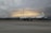 A large white aircraft with the inscription 'United States Dept. of Commerce' is parked on a tarmac. The sky in the background appears dramatic with layers of dark clouds and a faint glow of light, suggesting a sunrise or sunset.