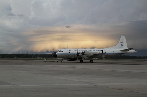 A large white aircraft with the inscription 'United States Dept. of Commerce' is parked on a tarmac. The sky in the background appears dramatic with layers of dark clouds and a faint glow of light, suggesting a sunrise or sunset.