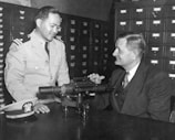 Two men are sitting at a table, one in a military uniform and the other in a suit. They are engaged in conversation, possibly discussing the optical instrument placed on the table. The background features several rows of filing cabinet drawers.