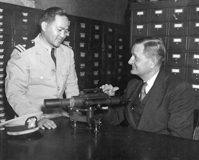 Two men are sitting at a table, one in a military uniform and the other in a suit. They are engaged in conversation, possibly discussing the optical instrument placed on the table. The background features several rows of filing cabinet drawers.