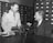 Two men are sitting at a table, one in a military uniform and the other in a suit. They are engaged in conversation, possibly discussing the optical instrument placed on the table. The background features several rows of filing cabinet drawers.