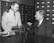 Two men are sitting at a table, one in a military uniform and the other in a suit. They are engaged in conversation, possibly discussing the optical instrument placed on the table. The background features several rows of filing cabinet drawers.