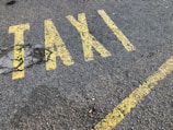 Yellow letters painted on a rough asphalt surface spell out the word 'TAXI'. The paint appears slightly worn and is accompanied by a yellow line running parallel to the word.