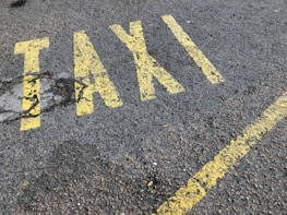Yellow letters painted on a rough asphalt surface spell out the word 'TAXI'. The paint appears slightly worn and is accompanied by a yellow line running parallel to the word.