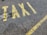 Yellow letters painted on a rough asphalt surface spell out the word 'TAXI'. The paint appears slightly worn and is accompanied by a yellow line running parallel to the word.