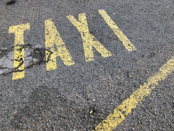 Yellow letters painted on a rough asphalt surface spell out the word 'TAXI'. The paint appears slightly worn and is accompanied by a yellow line running parallel to the word.