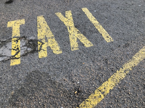 Yellow letters painted on a rough asphalt surface spell out the word 'TAXI'. The paint appears slightly worn and is accompanied by a yellow line running parallel to the word.