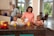 A family of four is gathered around a kitchen island. The mother is pouring orange juice from a carton into a glass for one of her young sons, while the other son watches with a smile. The father is standing by the countertop, holding a glass of juice. The kitchen is spacious with large windows and a variety of fruits displayed in a bowl.