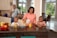 A family of four is gathered around a kitchen island. The mother is pouring orange juice from a carton into a glass for one of her young sons, while the other son watches with a smile. The father is standing by the countertop, holding a glass of juice. The kitchen is spacious with large windows and a variety of fruits displayed in a bowl.