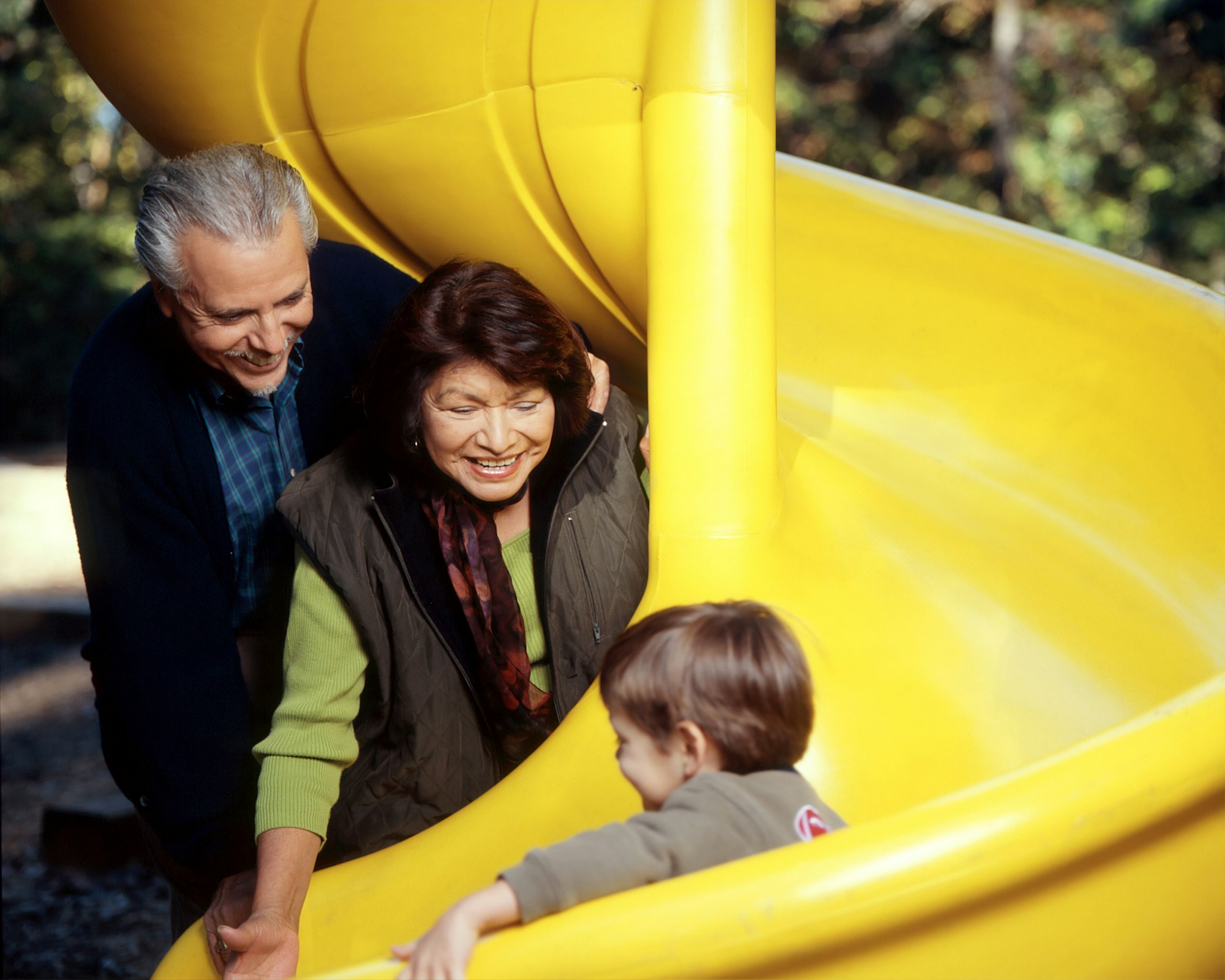 Family at a Playground. An older male and female standing at the bottom of a yellow slide while a young male child slides down. Photographer Rhoda Baer