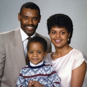 A family portrait featuring three people, consisting of two adults and a child. The man is wearing a beige suit and a tie, while the woman is dressed in a light pink top. The child is standing in front, wearing a patterned sweater. All appear to be smiling and are posed closely together in front of a neutral background.