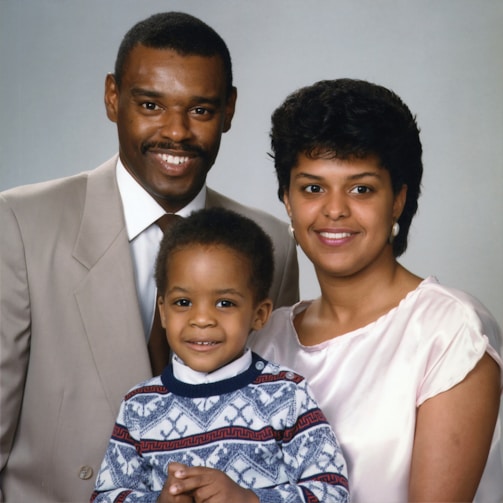 A family portrait featuring three people, consisting of two adults and a child. The man is wearing a beige suit and a tie, while the woman is dressed in a light pink top. The child is standing in front, wearing a patterned sweater. All appear to be smiling and are posed closely together in front of a neutral background.