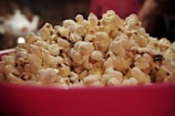 A large pile of fluffy, buttery popcorn fills a bright pink bowl, with some kernels having a golden hue. The background is blurred, focusing attention on the popcorn's texture and color.