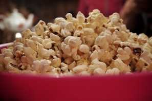 A large pile of fluffy, buttery popcorn fills a bright pink bowl, with some kernels having a golden hue. The background is blurred, focusing attention on the popcorn's texture and color.