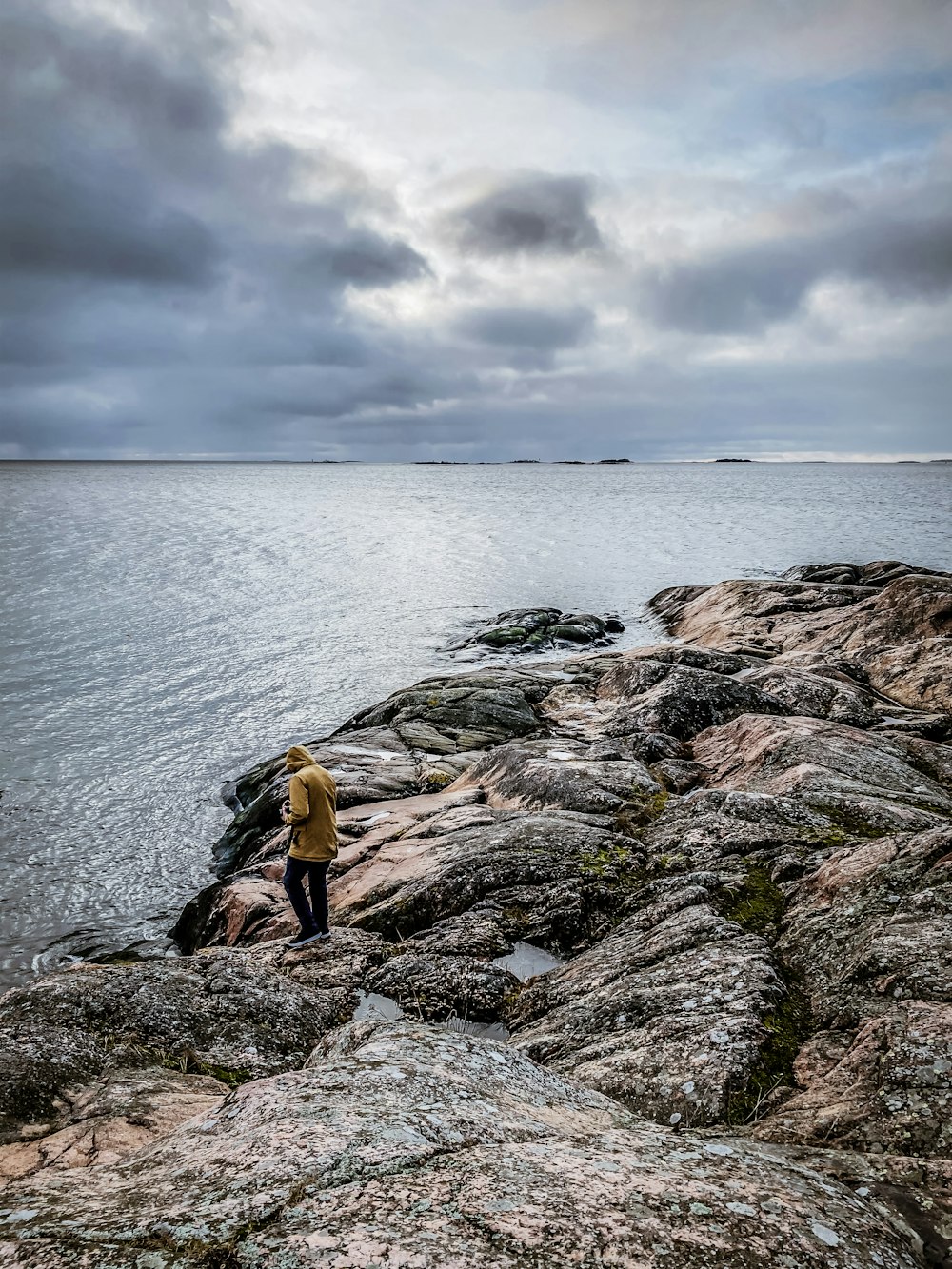 man wearing brown jacket standing on the stone near the body of water