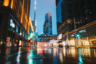 A vibrant cityscape at dusk, with glowing lights reflecting on wet streets under a dark sky.