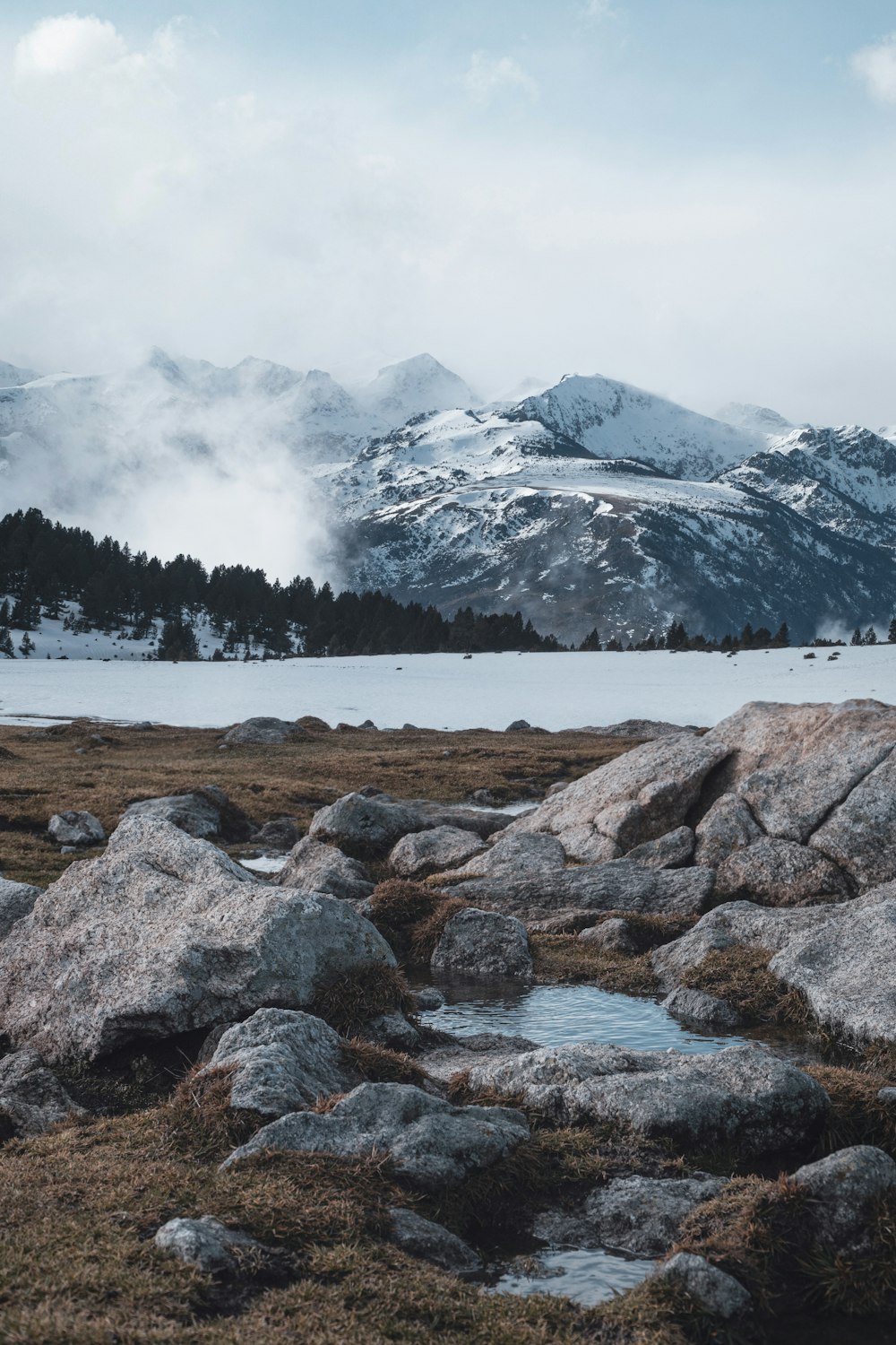 gray rock formations near body of water viewing mountain covered with snow under white and blue sky