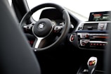 The interior of a car featuring a steering wheel with a logo at its center, dashboard controls, and a digital display. The dashboard includes climate control buttons and other vehicle control features.