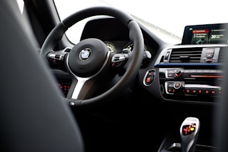 The interior of a car featuring a steering wheel with a logo at its center, dashboard controls, and a digital display. The dashboard includes climate control buttons and other vehicle control features.