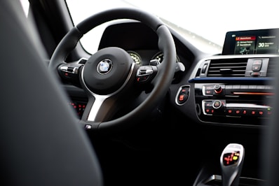 The interior of a car featuring a steering wheel with a logo at its center, dashboard controls, and a digital display. The dashboard includes climate control buttons and other vehicle control features.