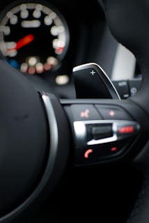 A close-up view of a car's steering wheel and control panel, focusing on a paddle shifter marked with a plus sign. The background shows a blurred speedometer with a red needle and illuminated controls.