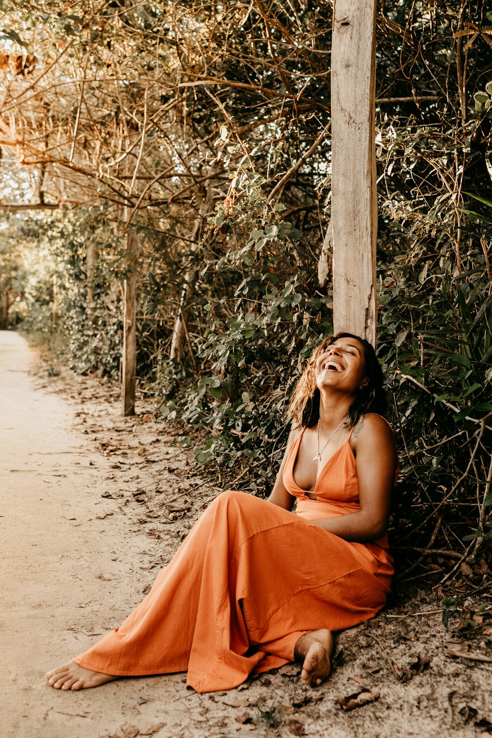 Barfüßige Frau im orangefarbenen Spaghettiträgerkleid lacht, während sie auf einem von grünen Bäumen umgebenen Weg sitzt