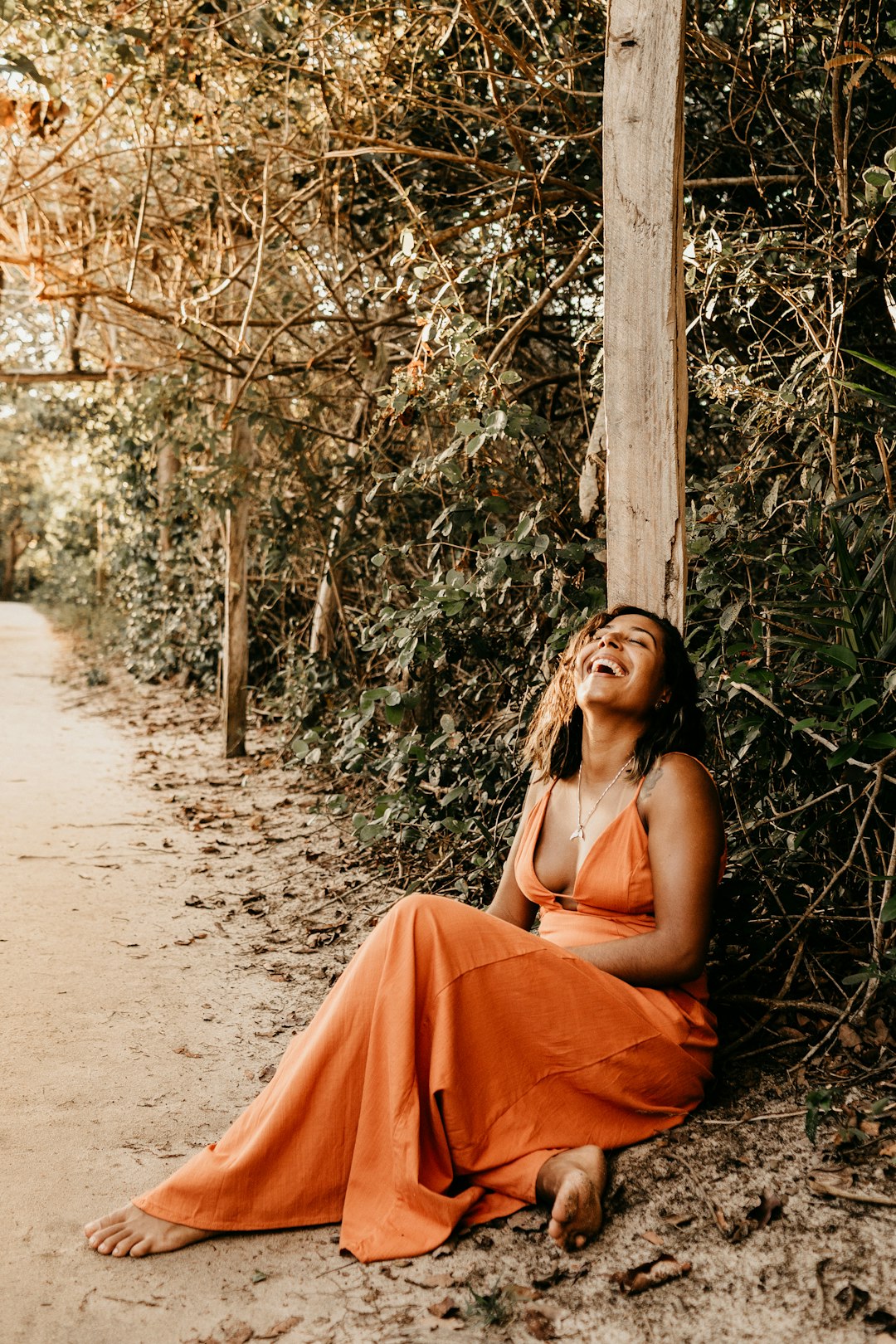 barefooted woman wearing orange spaghetti strap dress laughing while sitting on pathway surrounded with green trees