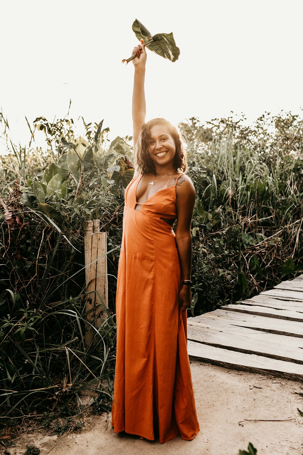 smiling woman wearing orange standing dress holding and raising green leaf while standing near plants