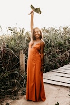 A woman in an orange dress stands on a path surrounded by lush greenery, holding up a leafy branch with a smile. Her pose suggests confidence and joy in a natural setting.