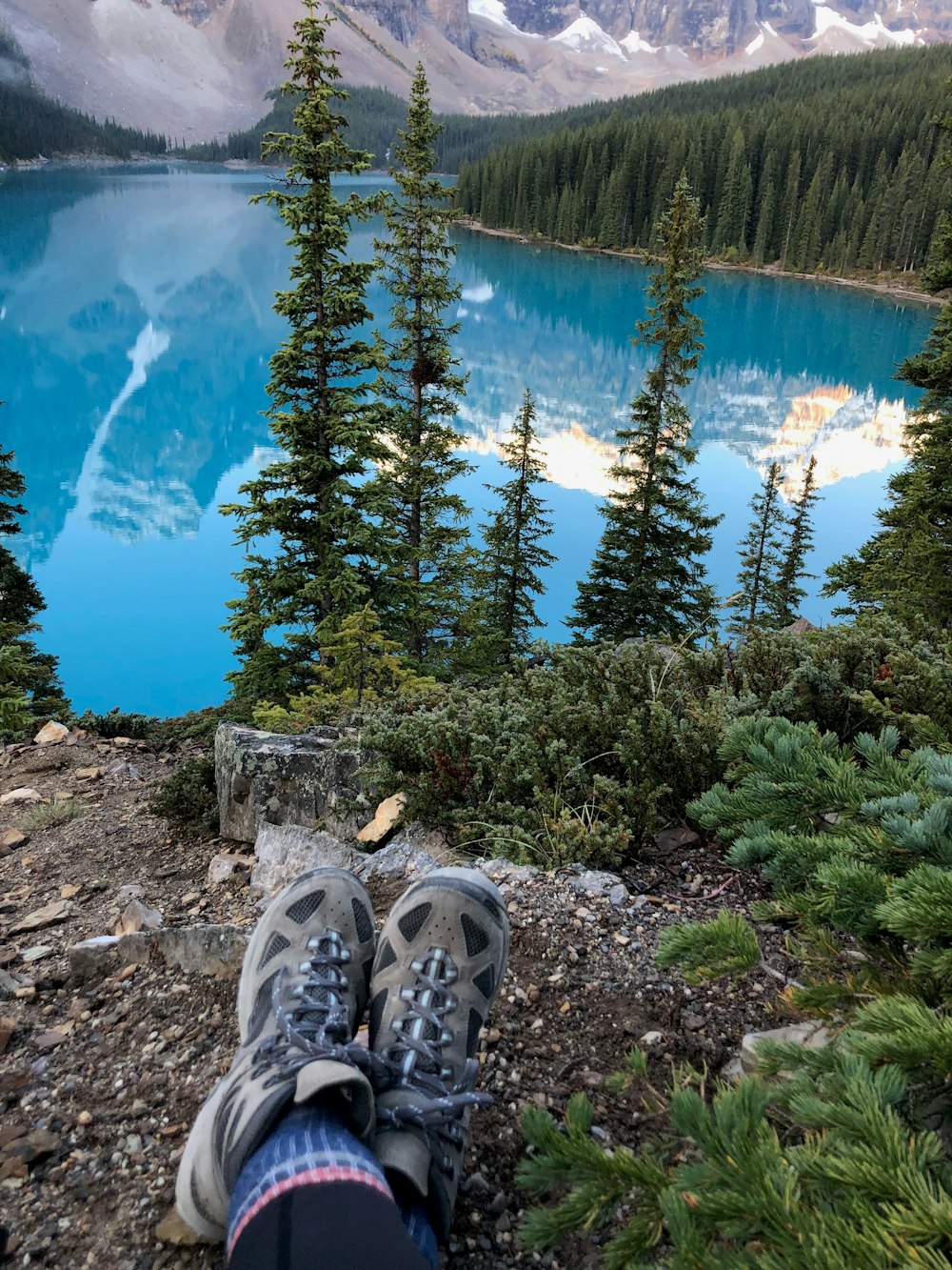 person sitting near lake surrounded with green trees viewing mountain during daytime