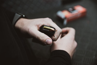 A close-up view of a person holding a magazine filled with bullets. The person's hands are visible, and they wear a dark sleeve. In the blurry background, there's an orange box of ammunition.