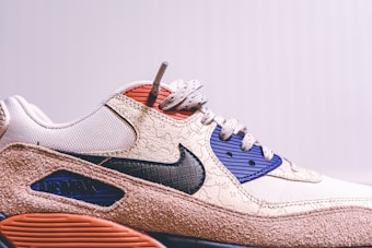 A close-up view of a stylish sneaker featuring a prominent swoosh logo on the side. The design includes textured patterns on a beige upper with navy and red details. The sole has a brown, rugged appearance.