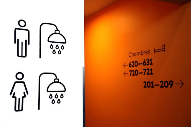 On the left, there are simplistic black line drawings of a male and female figure, each with a showerhead symbol above, indicating restroom or shower facilities. On the right, an orange wall features black text with the word 'Chambres' and directional arrows pointing to room numbers, suggesting this is a hallway in a hotel or lodging establishment.