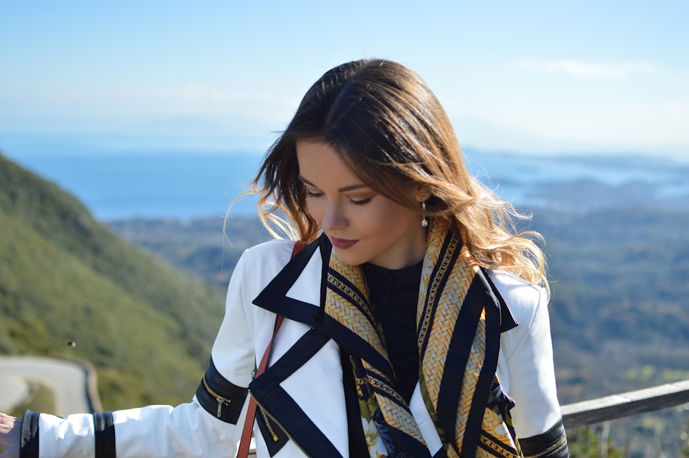 woman wearing white and black coat standing near railings while looking down viewing body of water and mountain under blue and white sky