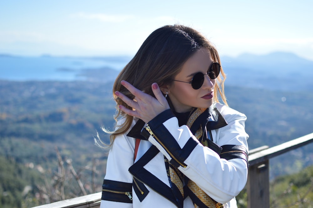 Mujer con abrigo blanco y negro y gafas de sol de pie mientras arregla su cabello viendo la montaña bajo el cielo azul y blanco