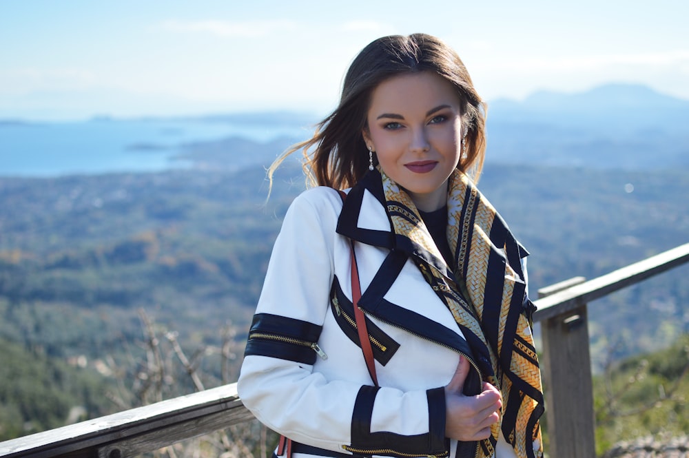 smiling woman wearing white and black coat standing near gray wooden railings viewing body of water and mountain under white and blue sky