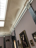 An art gallery with ornately decorated ceilings and walls showcasing various framed paintings. The gallery features classical architectural elements, including molding and pilasters. Several pieces of art, including portraits and larger paintings, hang on the walls.