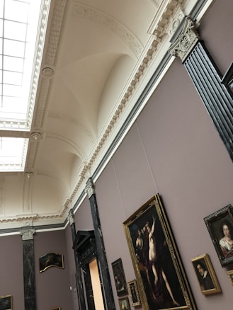 An art gallery with ornately decorated ceilings and walls showcasing various framed paintings. The gallery features classical architectural elements, including molding and pilasters. Several pieces of art, including portraits and larger paintings, hang on the walls.