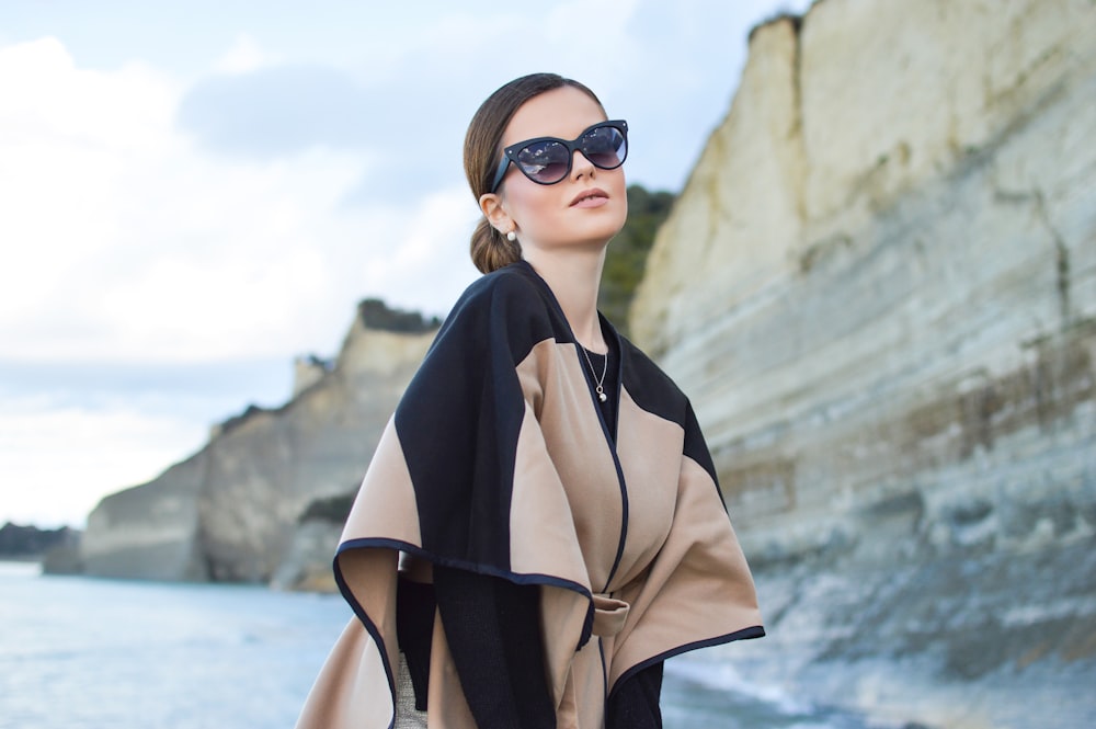 Frau in schwarz-braunem Kleid und Sonnenbrille steht in der Nähe eines Gewässers unter weißem und blauem Himmel