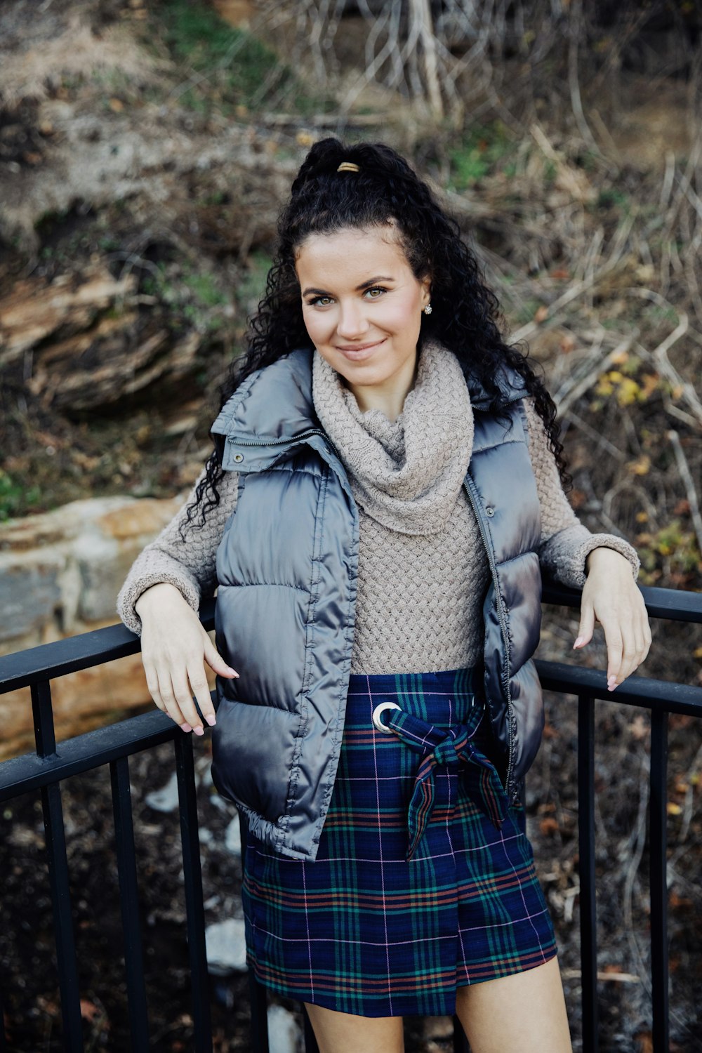 smiling woman wearing gray cowl-neck sweater, gray zip-up vest, and blue, red, and green plaid mini skirt standing and leaning on black metal railings
