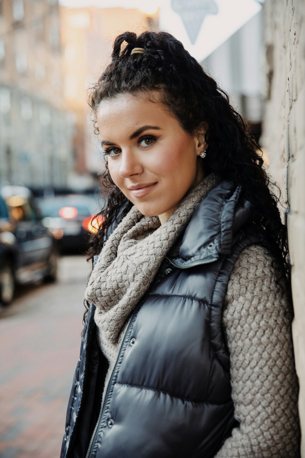 smiling woman wearing gray sweater and gray zip-up vest standing and leaning against the wall