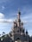 A fairytale castle with a pink and blue facade, featuring ornate spires and turrets with golden accents. The structure is situated on a rocky landscape with manicured trees nearby. A blue sky with scattered clouds is in the background.