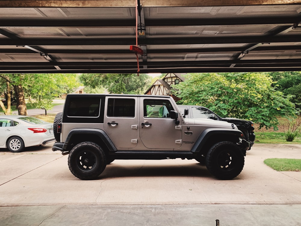 parked gray Jeep Wrangler SUV near white car, trees, and garage